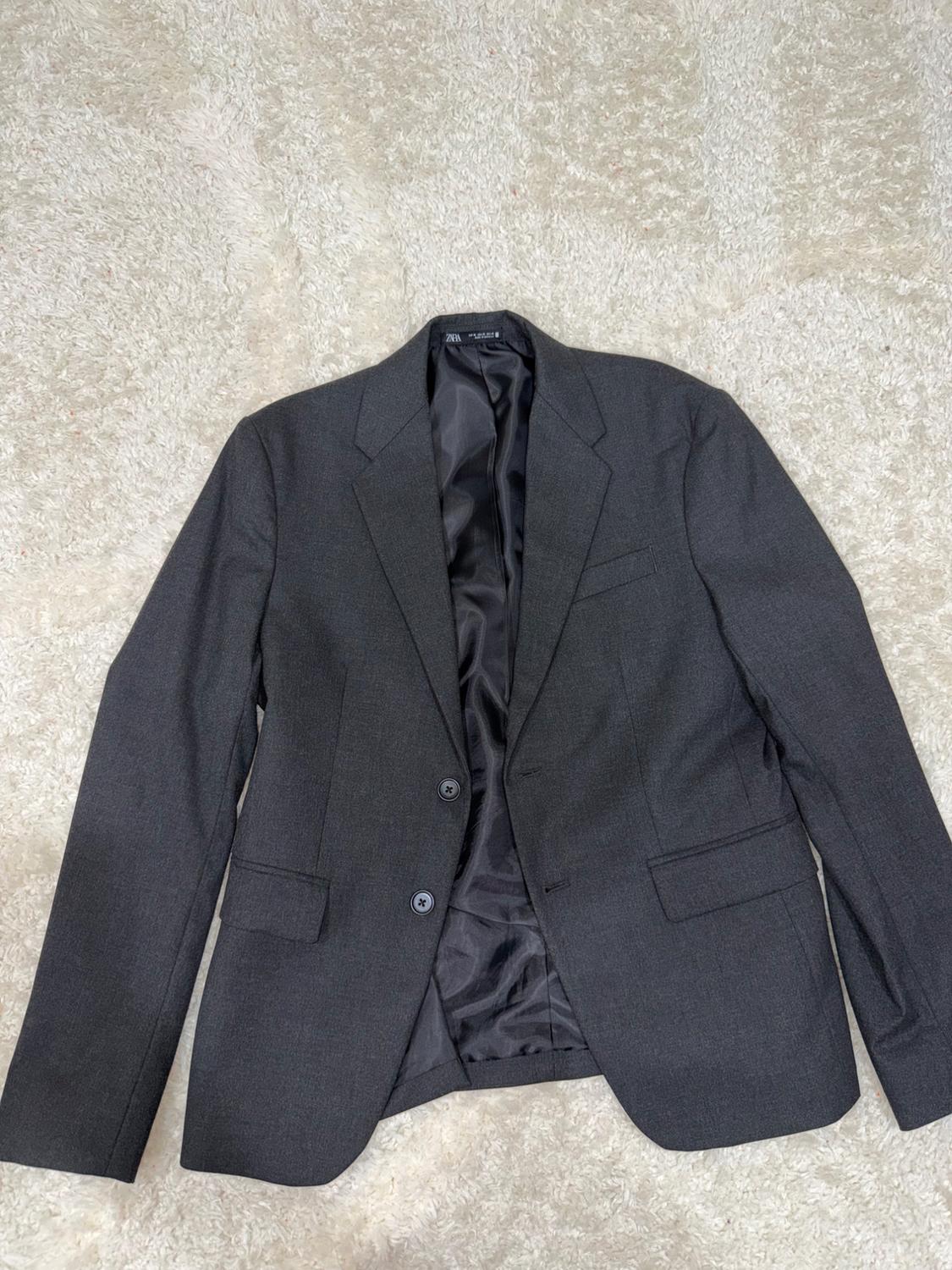 Dark grey two-button blazer with a ZARA label, open to show the black lining, laid out on a textured white carpet.
