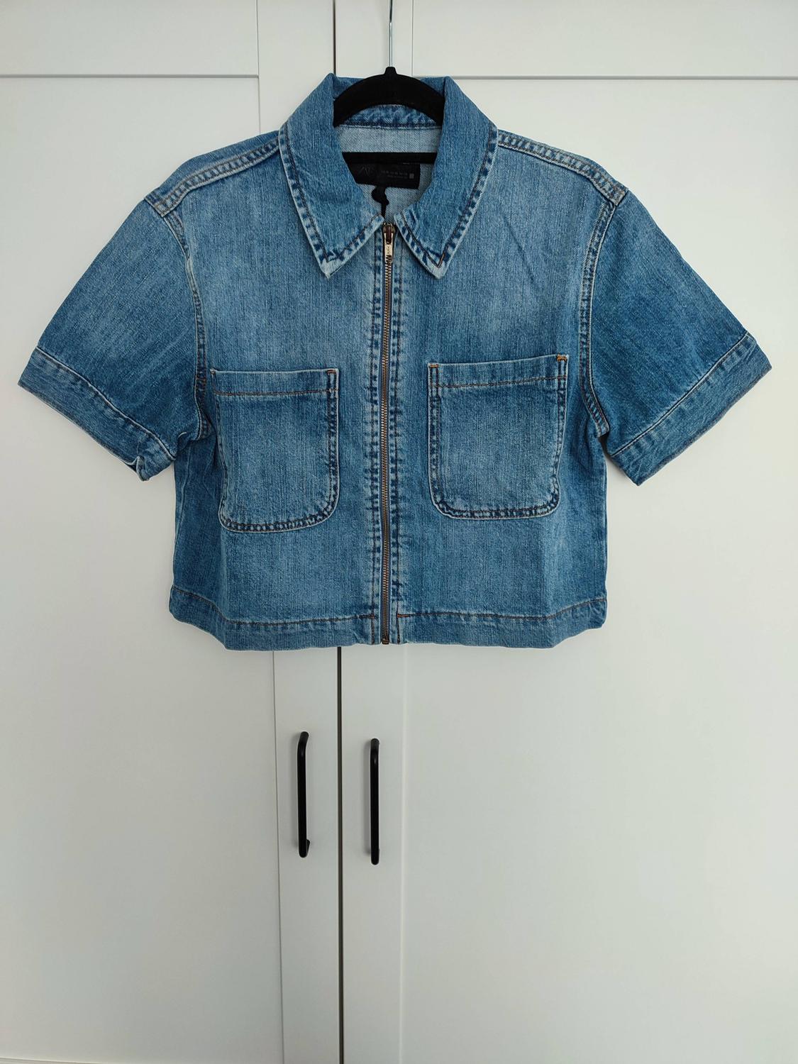 Short-sleeved blue denim shirt with a zip and front pockets, hanging on a black hanger against a white wardrobe.