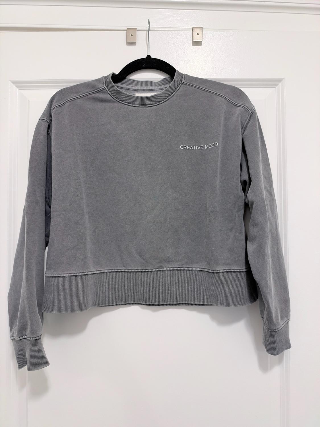 Gray long-sleeved sweatshirt hanging on a hanger over a white door. It has the text "CREATIVE MOOD" on the chest.