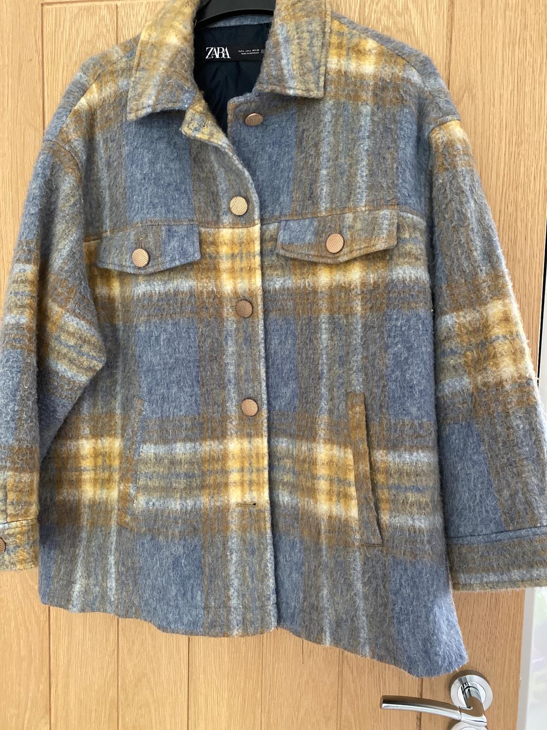 Grey and yellow checked jacket hanging on a wooden door. Brown buttons and front pockets. Visible label.