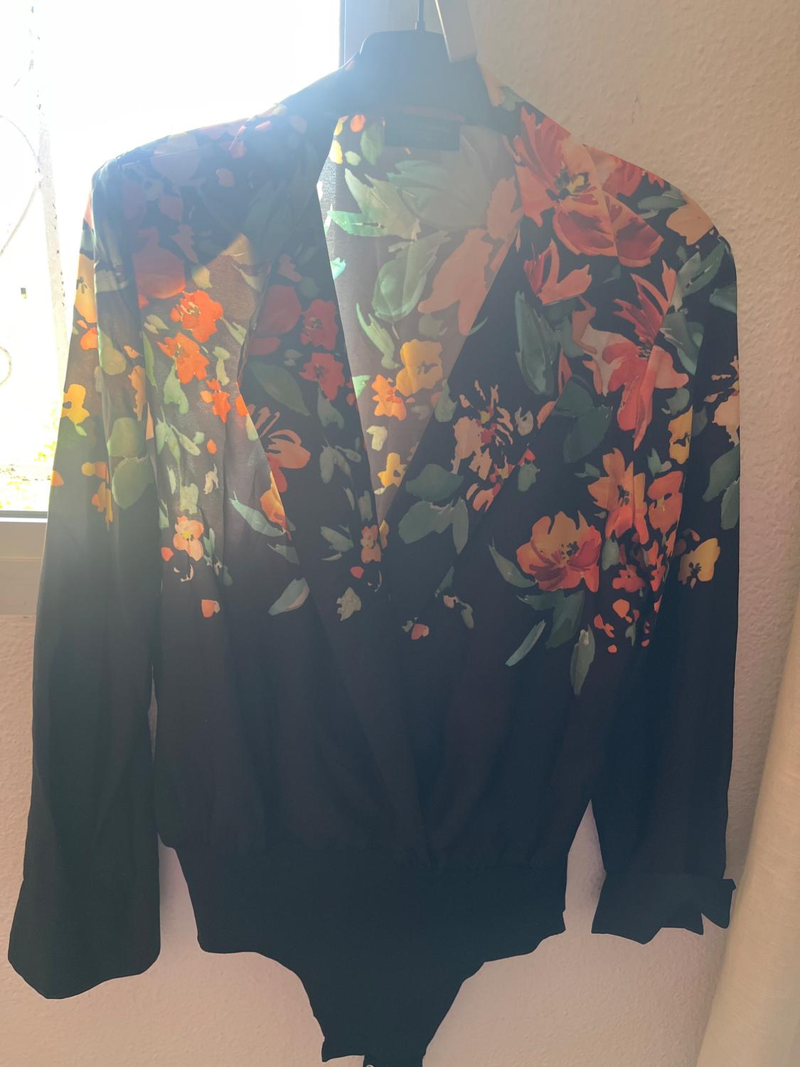 Long-sleeved blouse with a floral print in orange, yellow, and green tones, hanging on a hanger in front of a window.