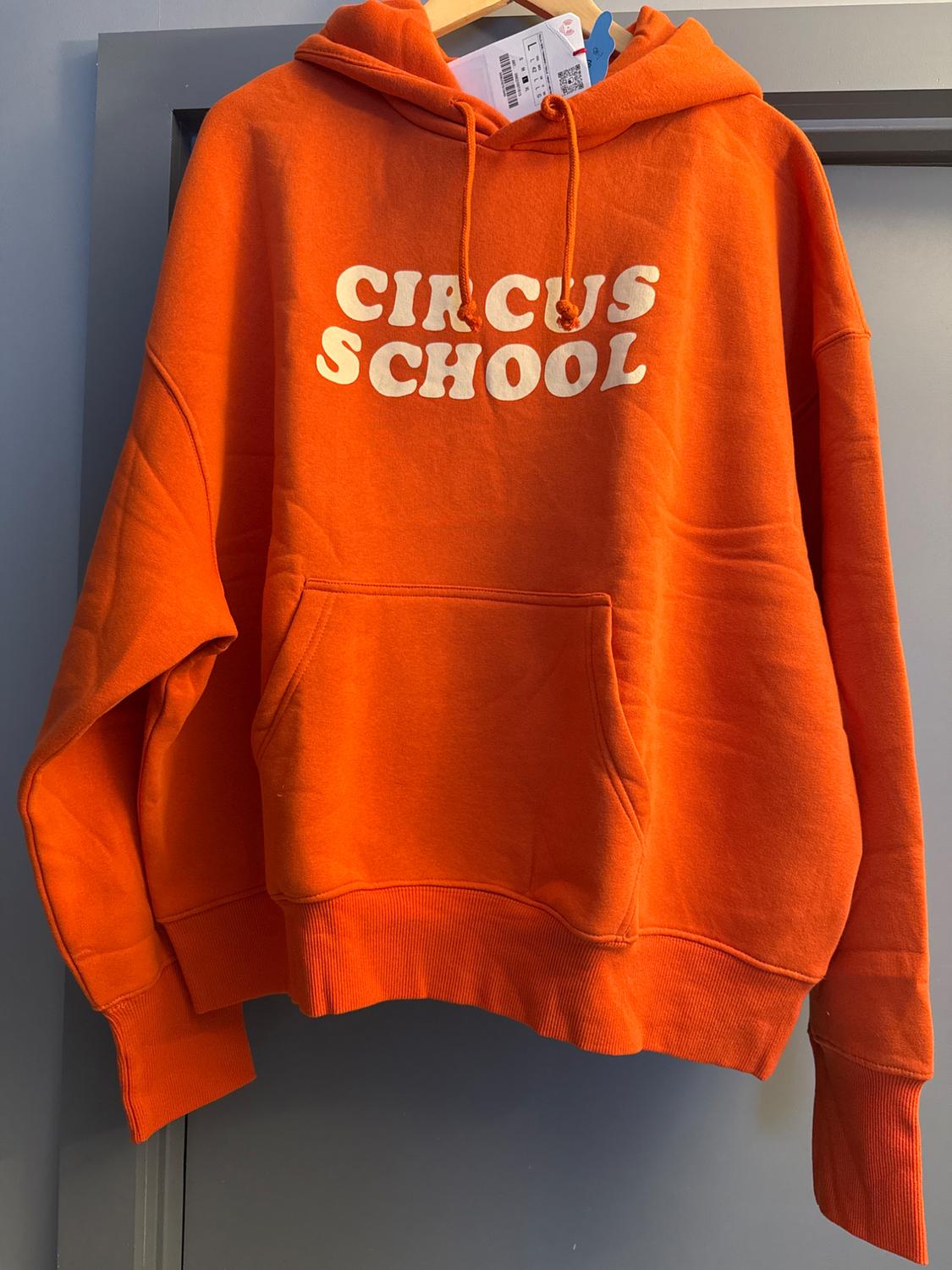 Orange hooded sweatshirt with a front pocket, hanging on a coat hanger. Text "CIRCUS SCHOOL" in white letters on the chest.