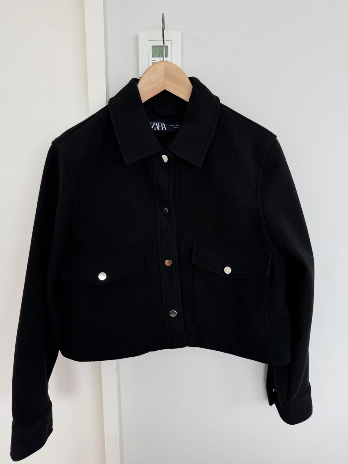 Black jacket hanging on a wooden hanger, with front pockets and silver buttons. Visible label with text "ZARA". White background.