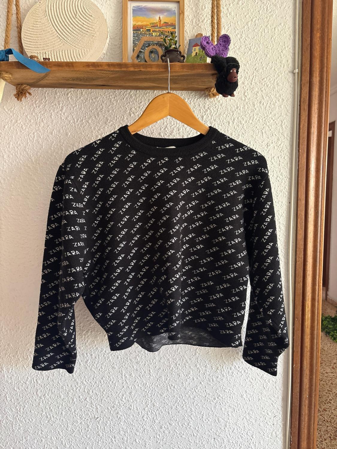Black jumper with a repeated white ZARA text print, hanging on a wooden hanger beneath a decorated shelf.