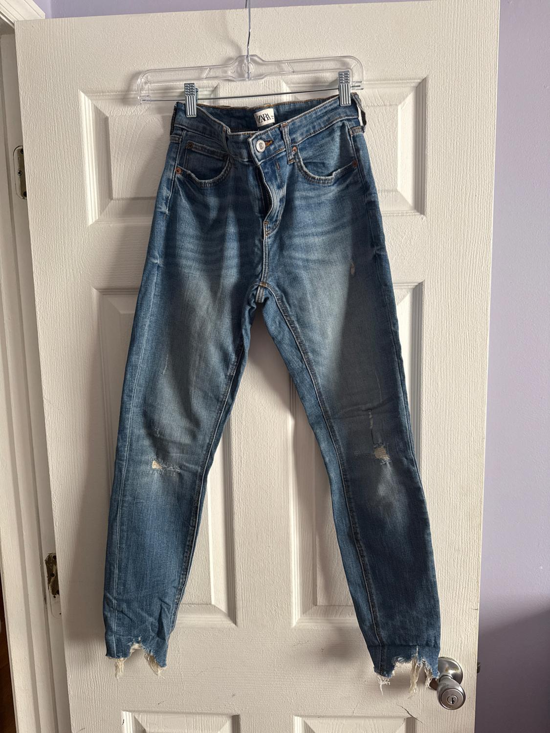 Distressed blue jeans hanging on a clear hanger over a white door. The label reads "Zara".