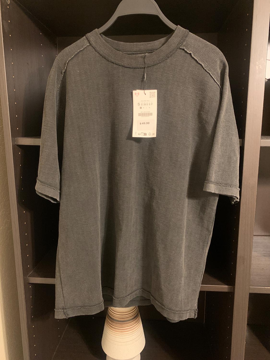 Grey short-sleeved t-shirt hanging on a black hanger, with a price tag of $49.90, on a dark wooden shelf.