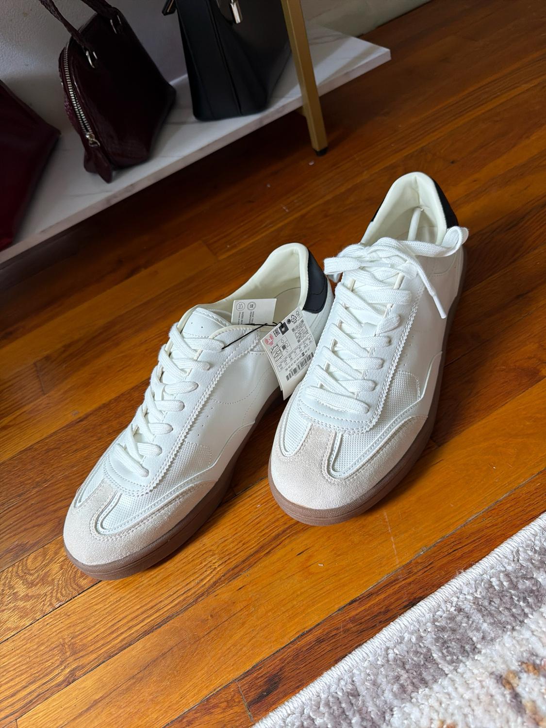 White trainers with a black heel, suede detailing and a brown sole on a wooden floor. In the background, a shelf with handbags.
