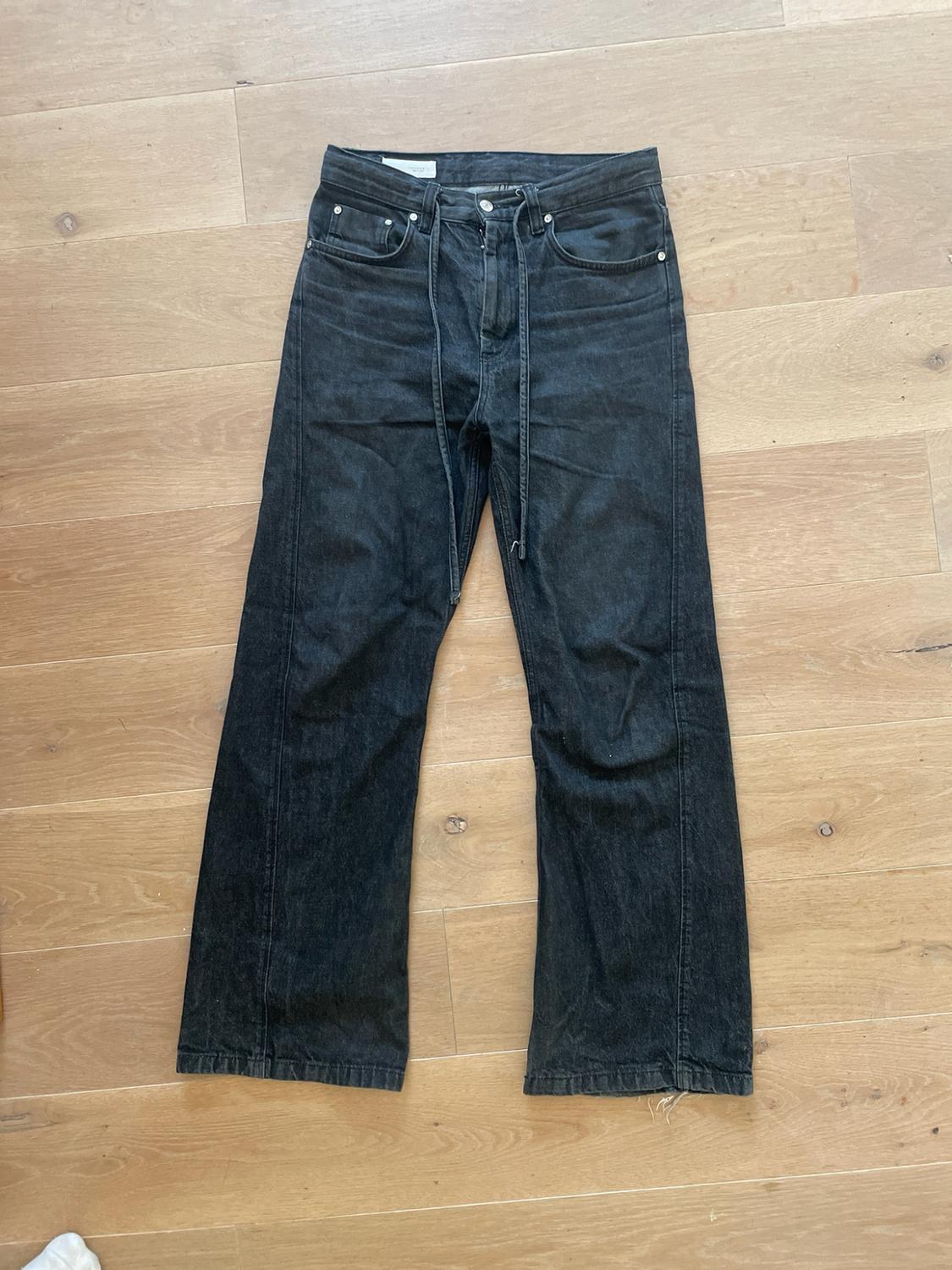 Dark grey denim trousers with front seams and a drawstring waist, laid out on a light wooden floor.