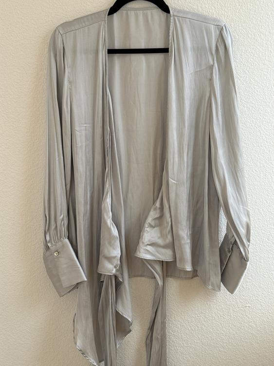 Light gray long-sleeved blouse with wide cuffs and ties, hanging on a black hanger against a white wall.