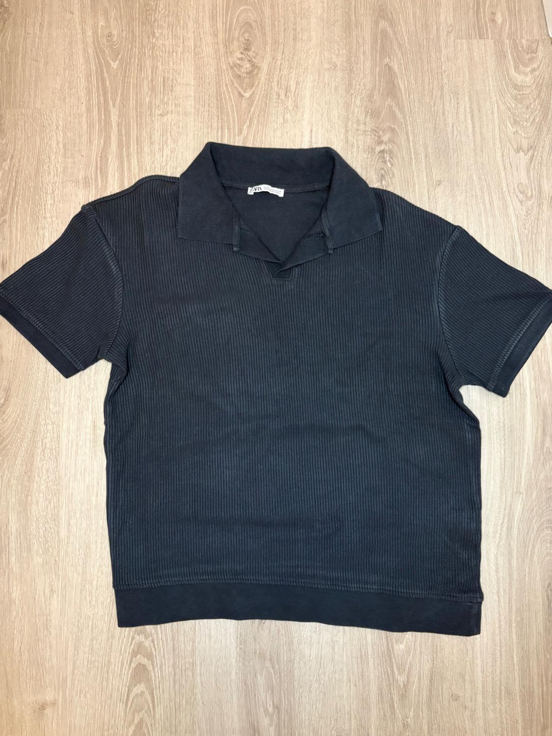 Black short-sleeved ribbed polo shirt on a light wood background. Visible label on the collar.