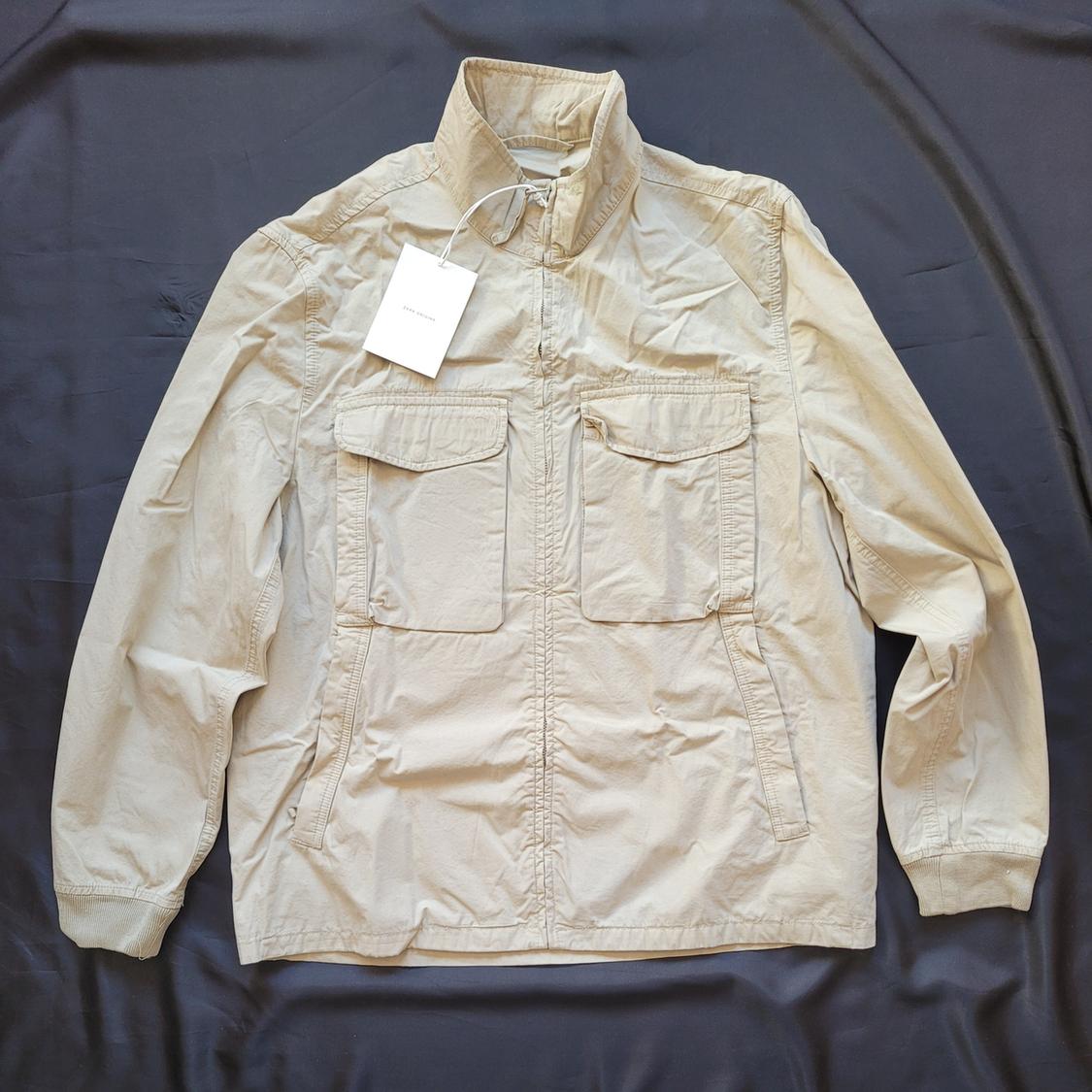 Beige jacket with a high collar and front pockets on a black background. White tag hanging from the collar.