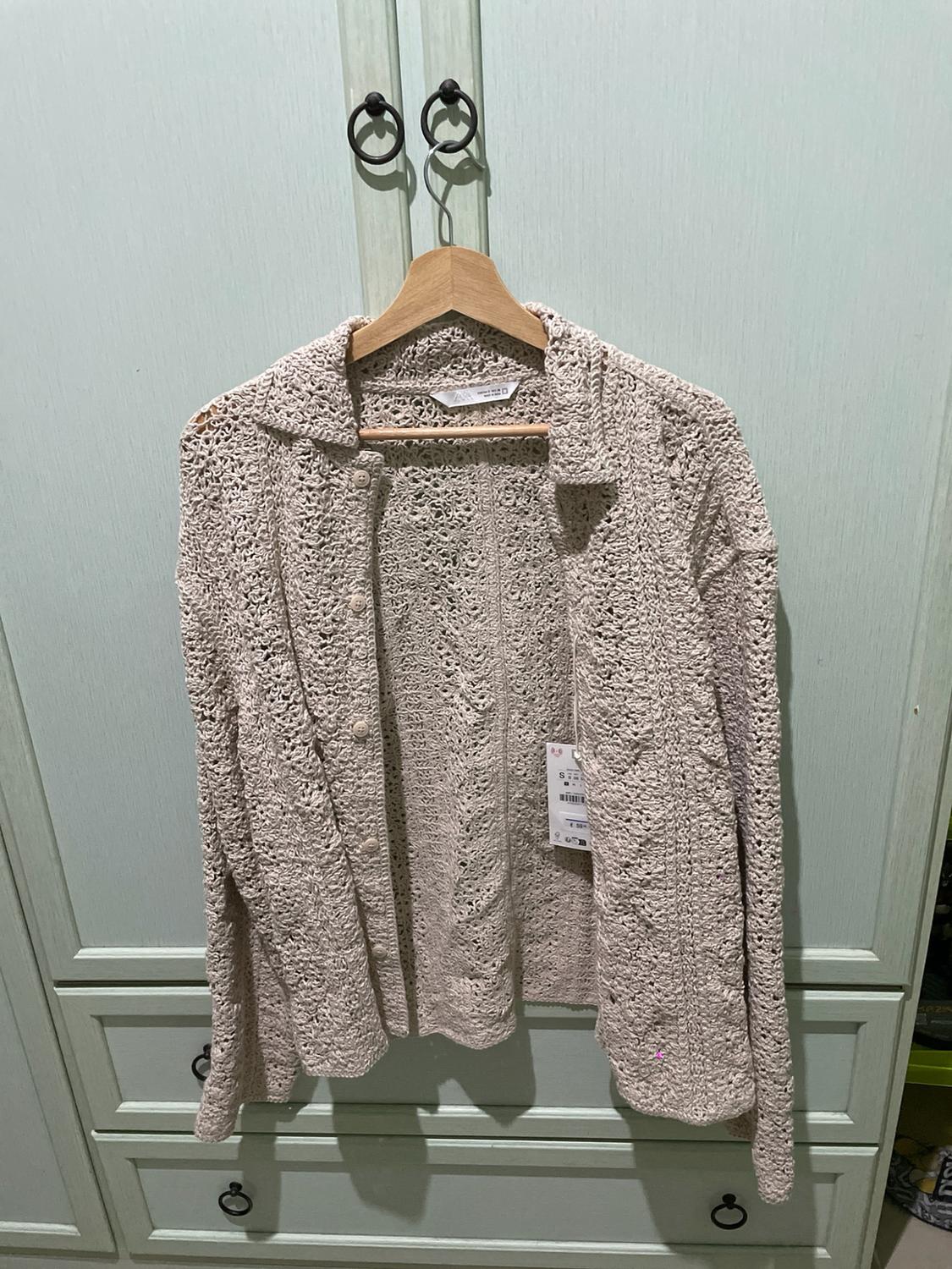 Beige knitted button-down shirt, hanging on a wooden coat hanger in front of a pastel green wardrobe.