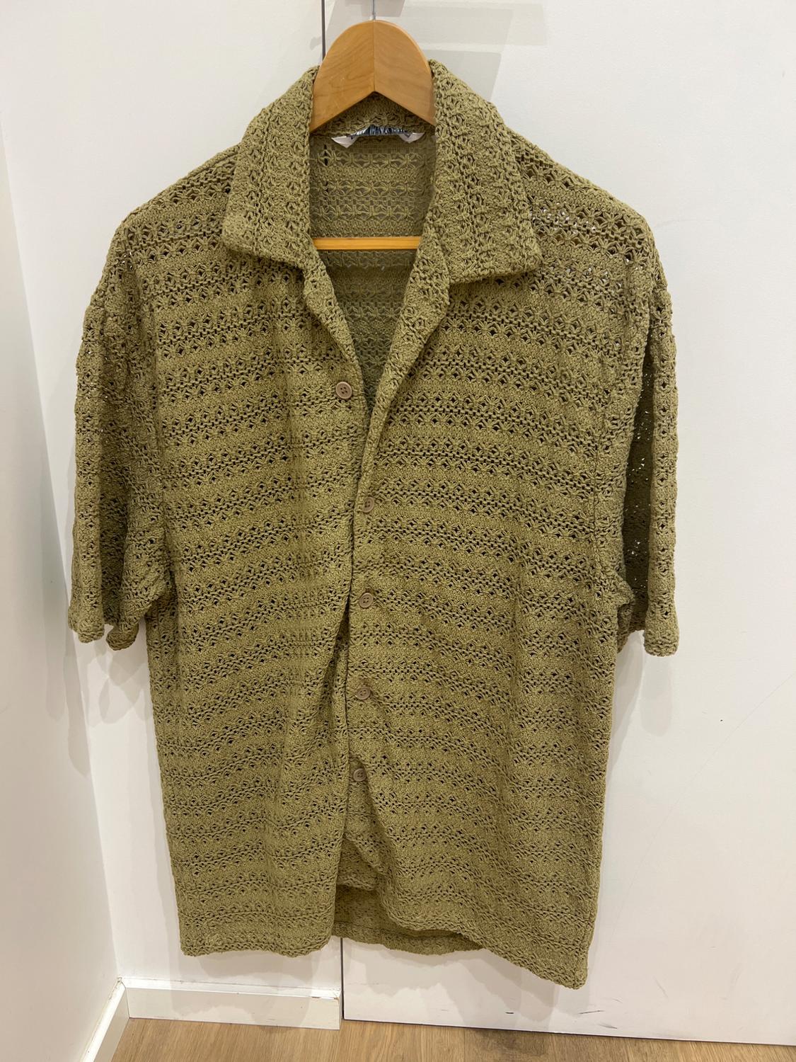 Olive green crochet shirt with buttons, hanging on a wooden coat hanger against a white wall.