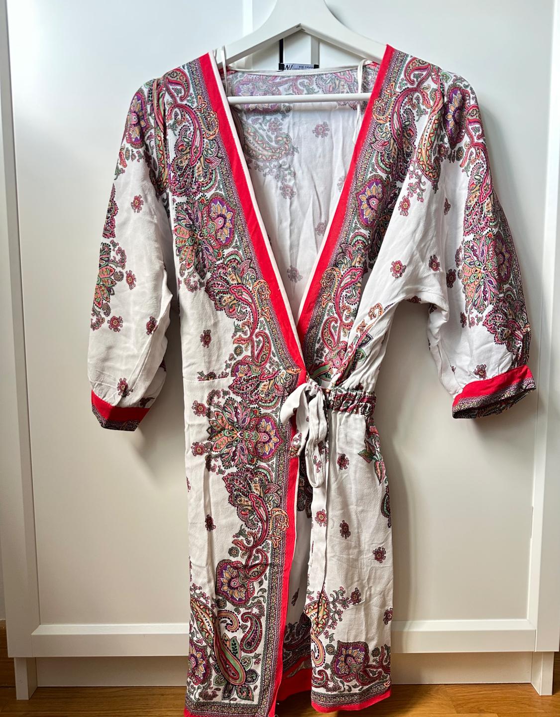 White wrap dress with multicoloured print, pink trim and waist tie, hanging on a hanger against a white door.