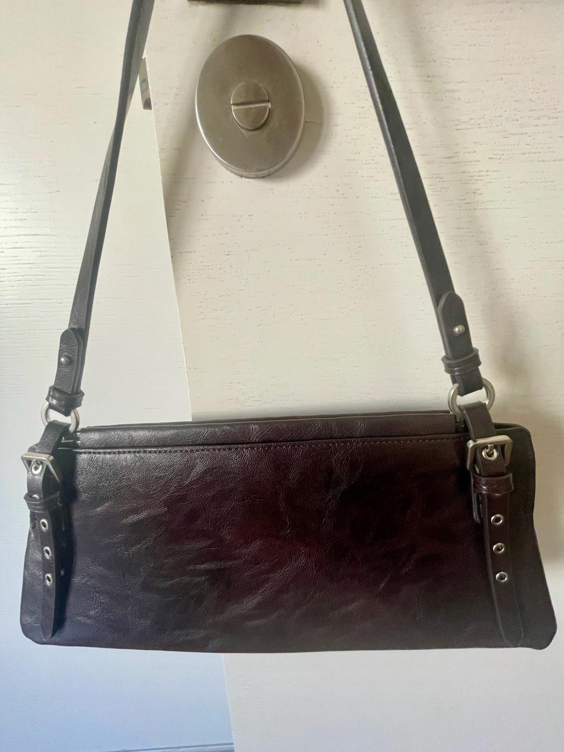 Dark brown rectangular shoulder bag hanging from the knob of a white door. Its texture and the straps with silver buckles stand out.