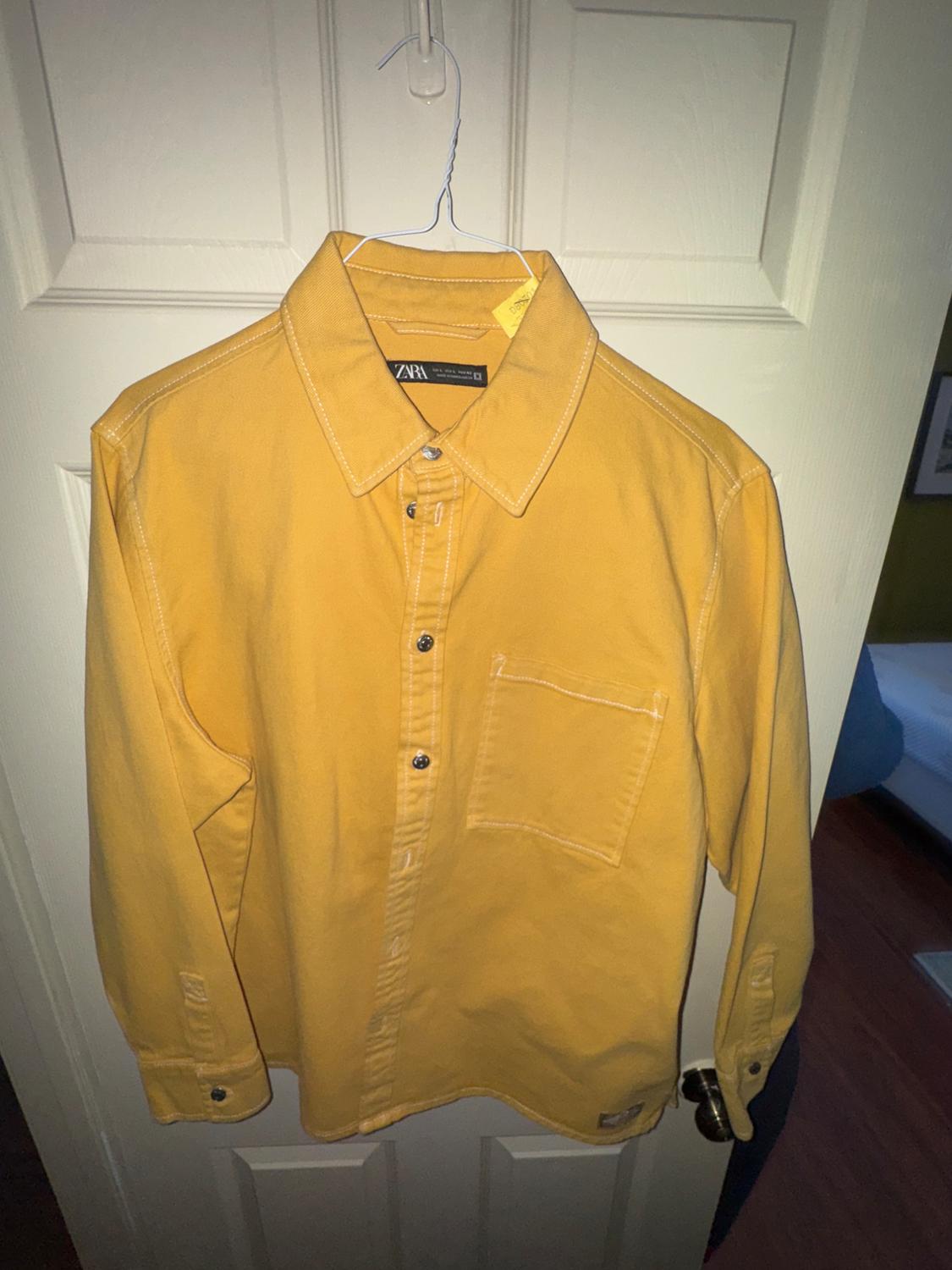 Yellow shirt hanging on a white hanger over a white door. The label says "ZARA". The shirt has a chest pocket.