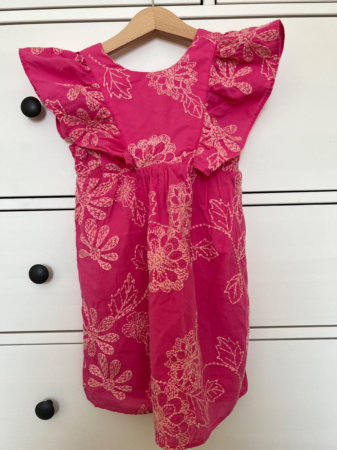 Pink dress with white floral embroidery, ruffled sleeves, hanging on a wooden hanger in front of a white chest of drawers.