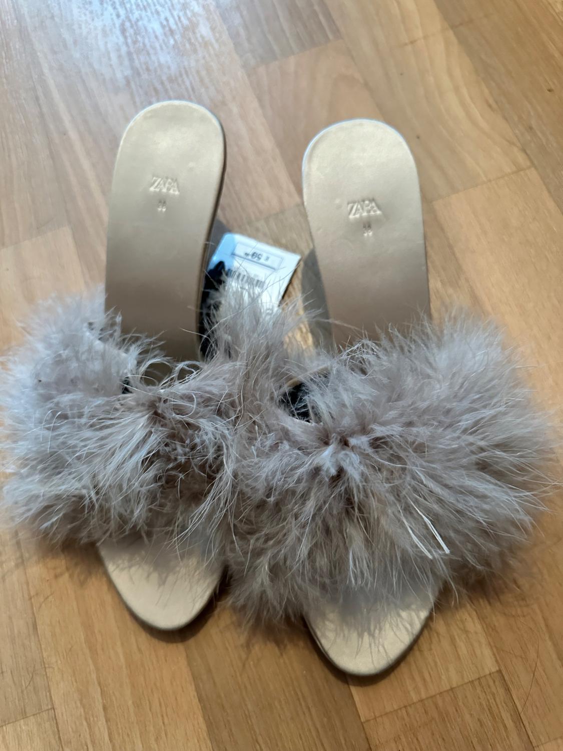 Beige sandals with grey feather detailing on the upper, placed on a light wood floor. Label visible.