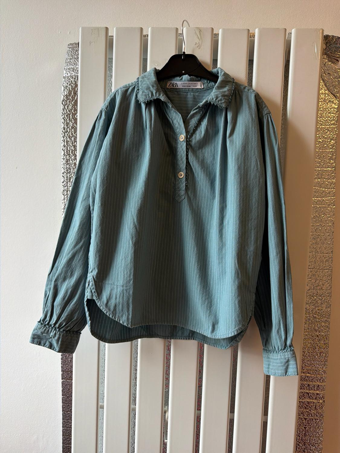 Long-sleeved blue shirt with white stripes and a zara label, hanging on a white radiator against a silver background.