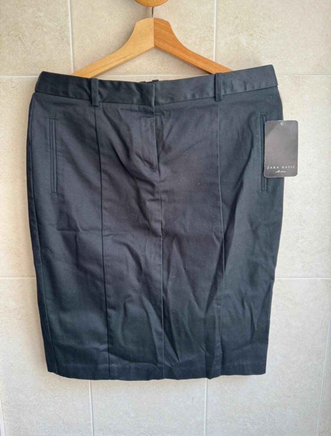 Black fabric skirt hanging on a wooden hanger, with a visible label that reads "ZARA BASIC". Beige tiled background.