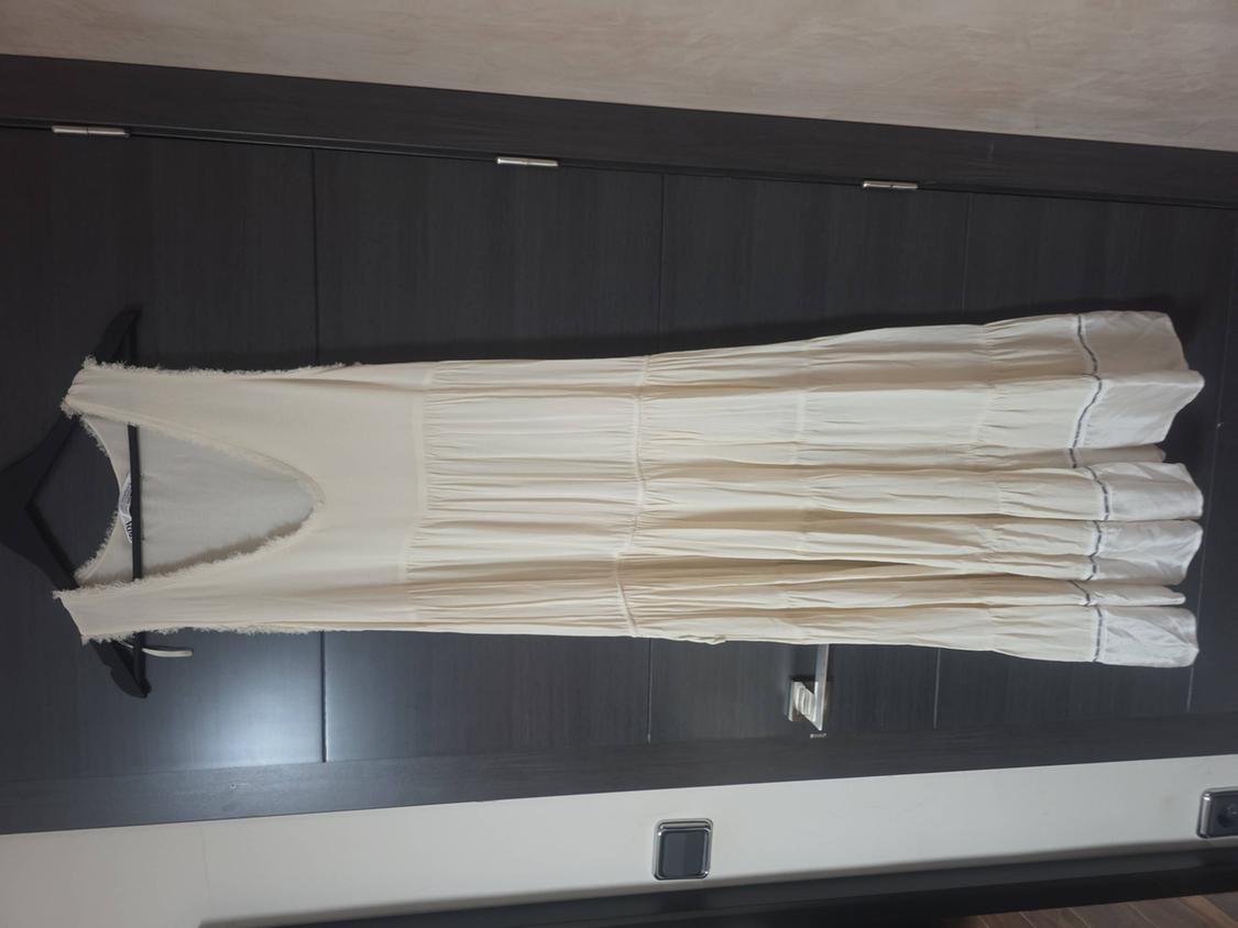 Cream-coloured sleeveless maxi dress with a frayed V-neck and tiered skirt, hanging on a hanger against a dark wood wardrobe.