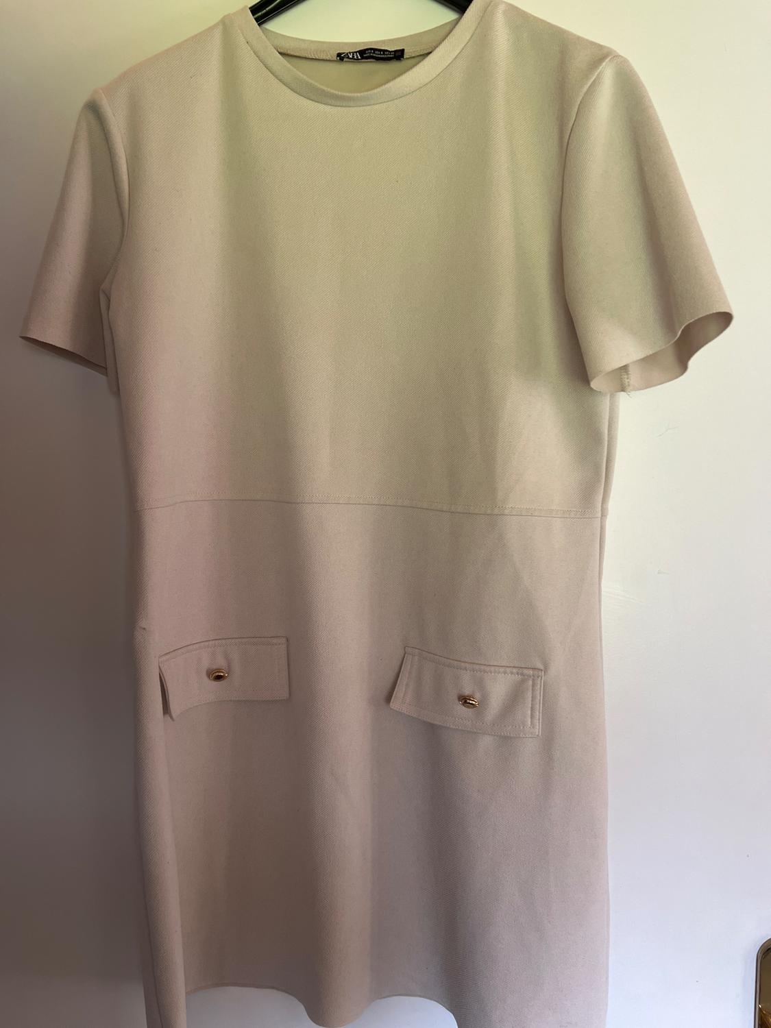 Beige short-sleeved dress on a black hanger. Round neck, seam at the waist and two front pockets with gold buttons.