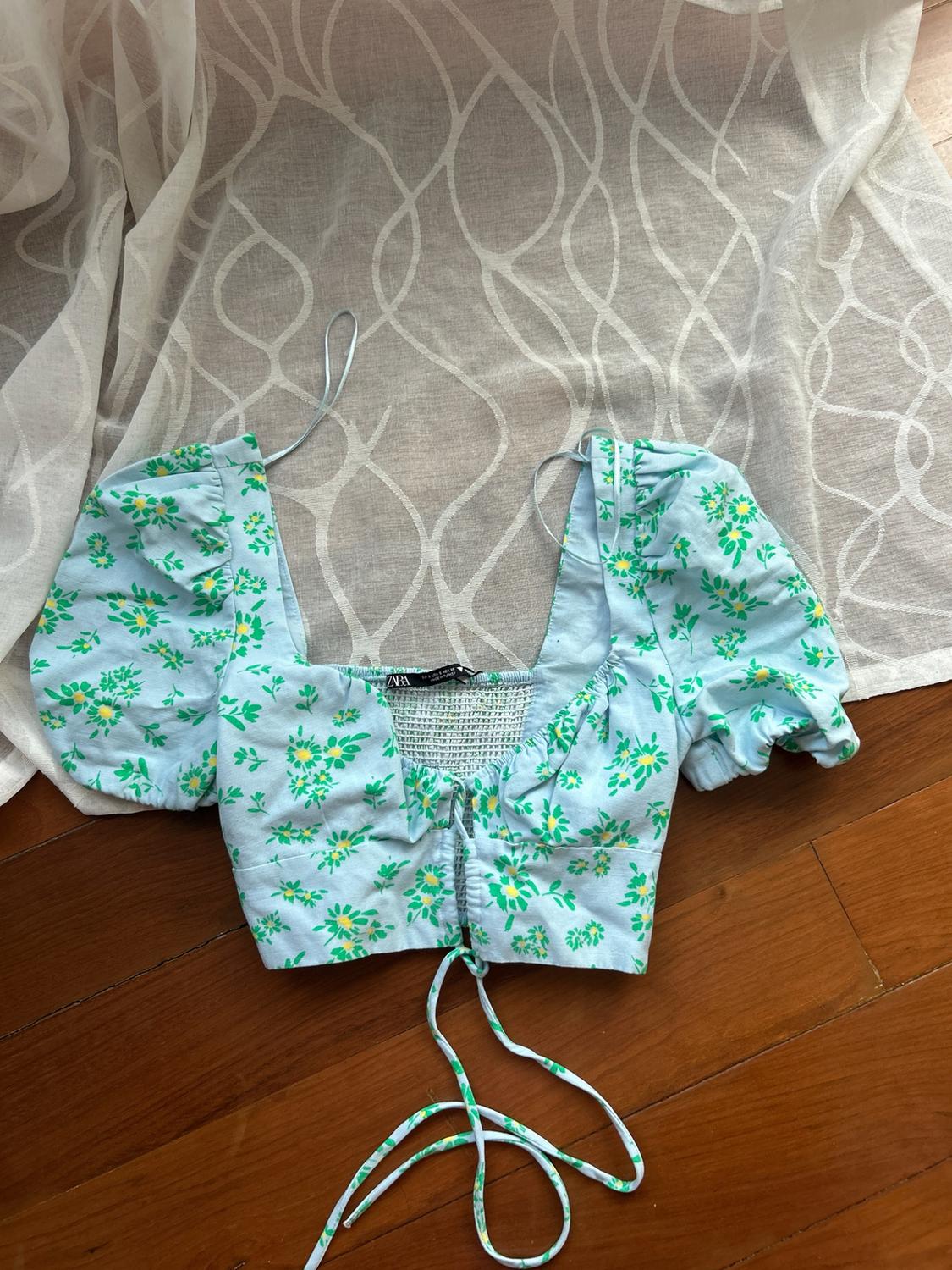 Short blue top with a green floral print, puff sleeves and a front tie, on a wooden floor and white fabric. Text: ZARA.
