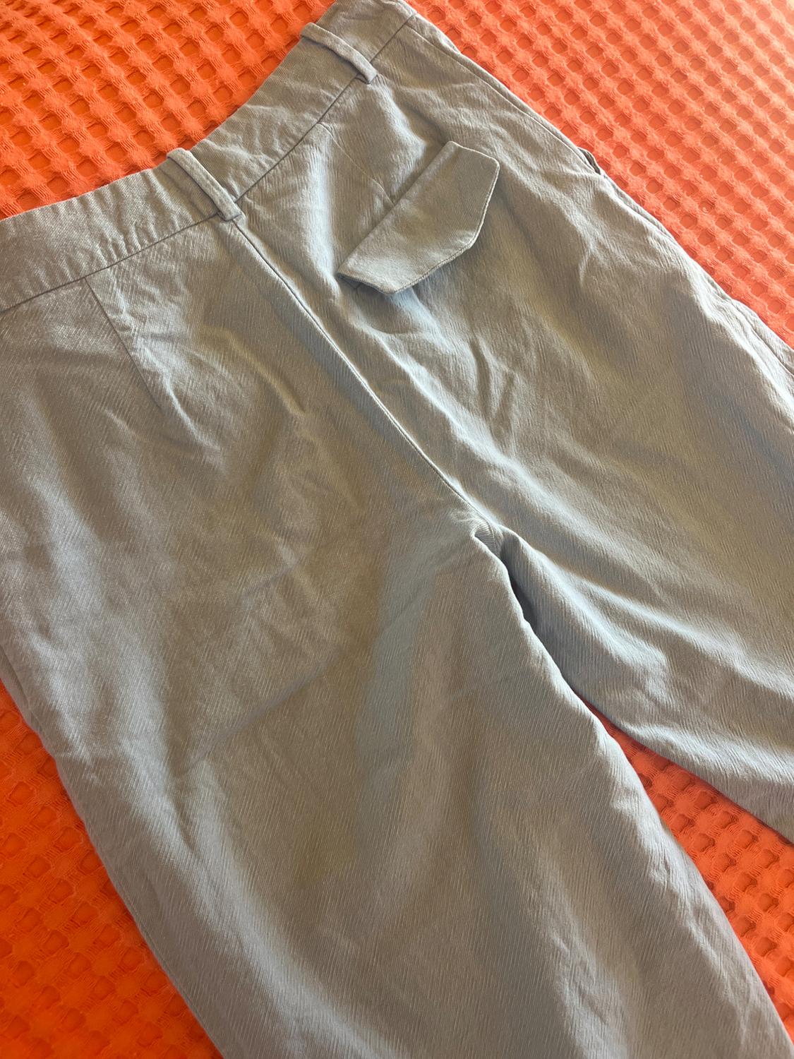 Light blue trousers with a flap back pocket and belt loops, laid out on a textured orange fabric. Casual style.