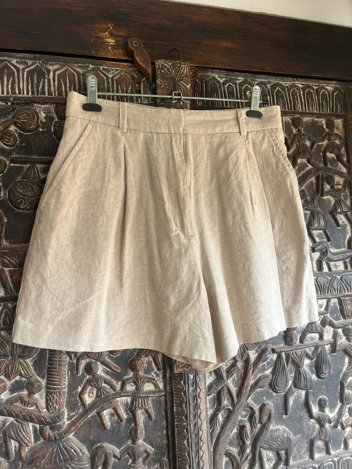 Beige pleated shorts hanging on a hanger against a dark, carved wooden background featuring figures. Relaxed style.