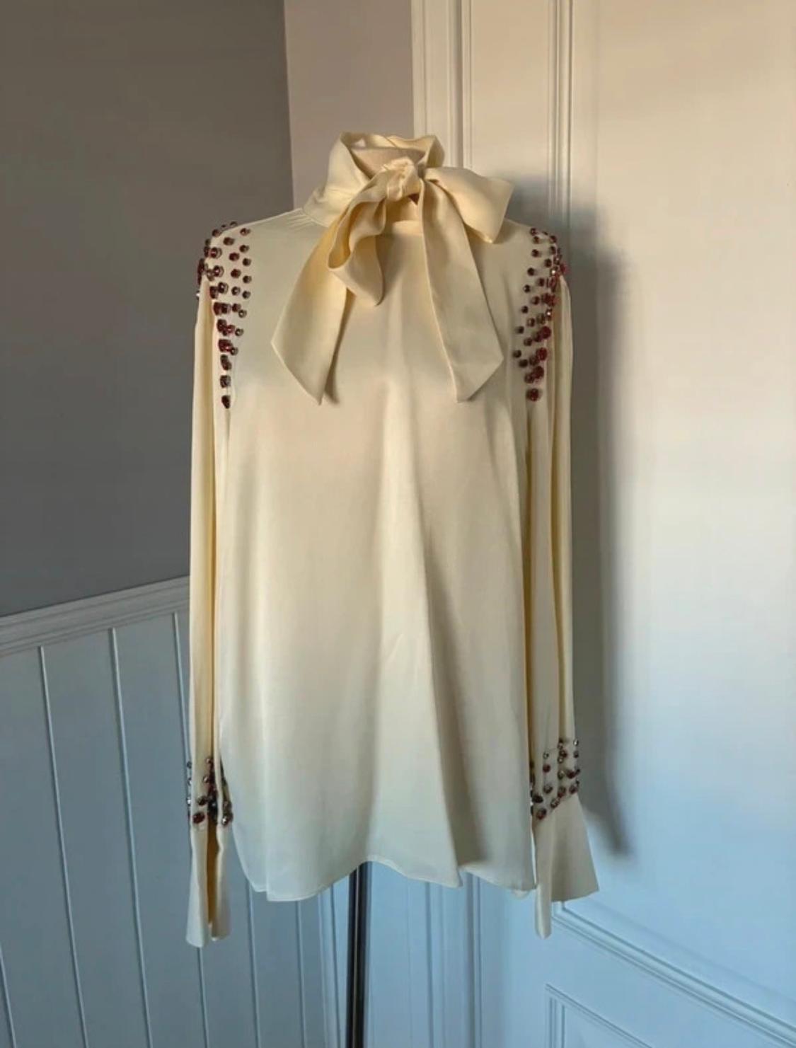 Cream blouse with a bow at the neck and diamante details on the shoulders and sleeves, hanging on a mannequin in front of a white wall.