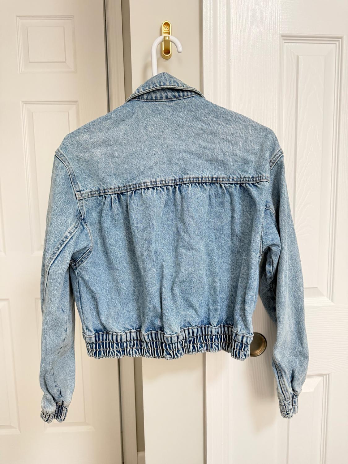 Light blue denim jacket seen from the back, hanging on a white door. Its gathered details on the back, waist, and cuffs stand out.