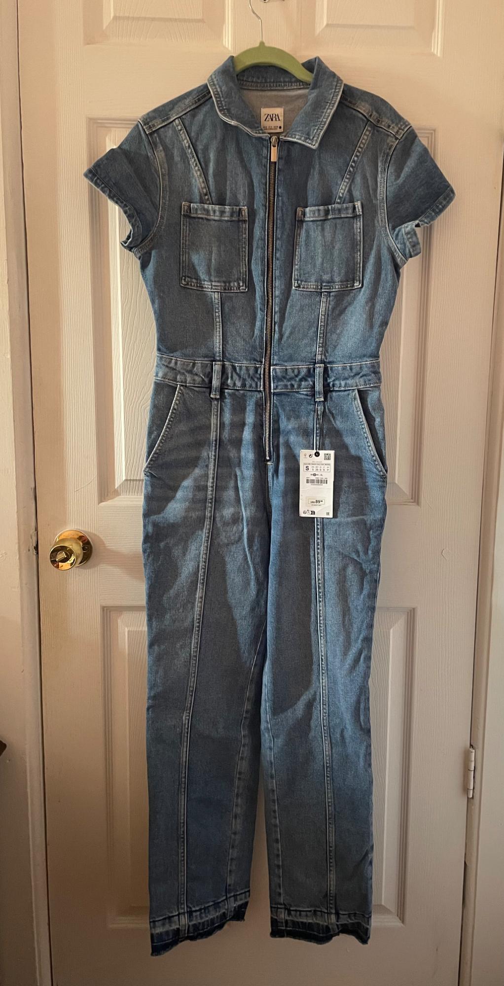 Blue denim jumpsuit with front zip and pockets, hanging on a white door. Visible tag with product details.