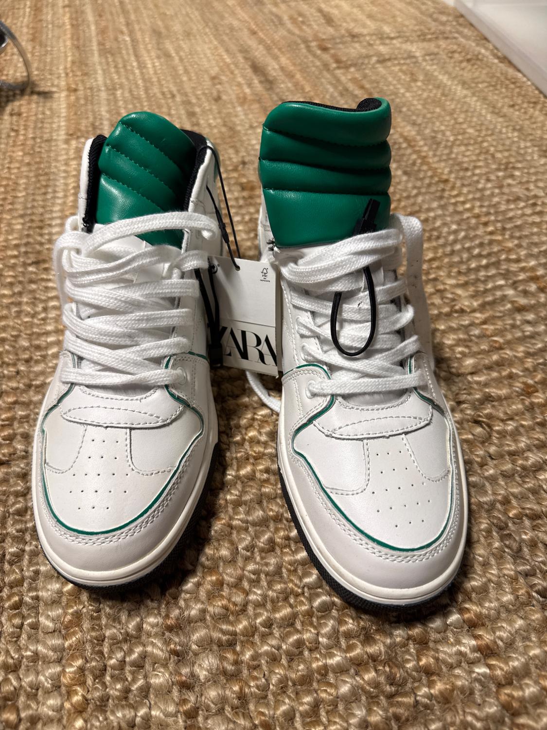 White trainers with green details and white laces on a woven rug. Visible label with text "ZARA".