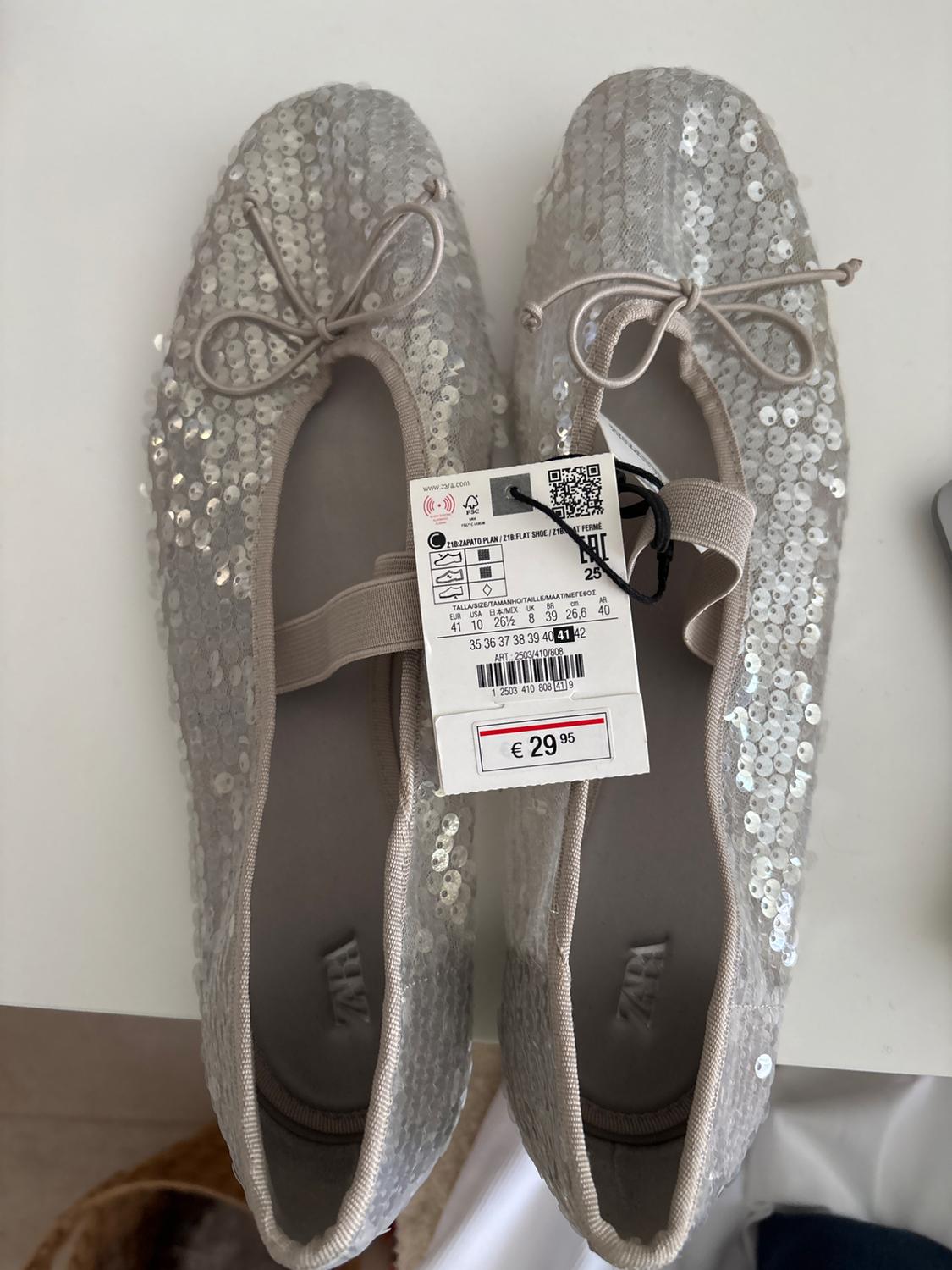 Silver sequin flat shoes with a bow, price tag of €29.95. White background, top view.