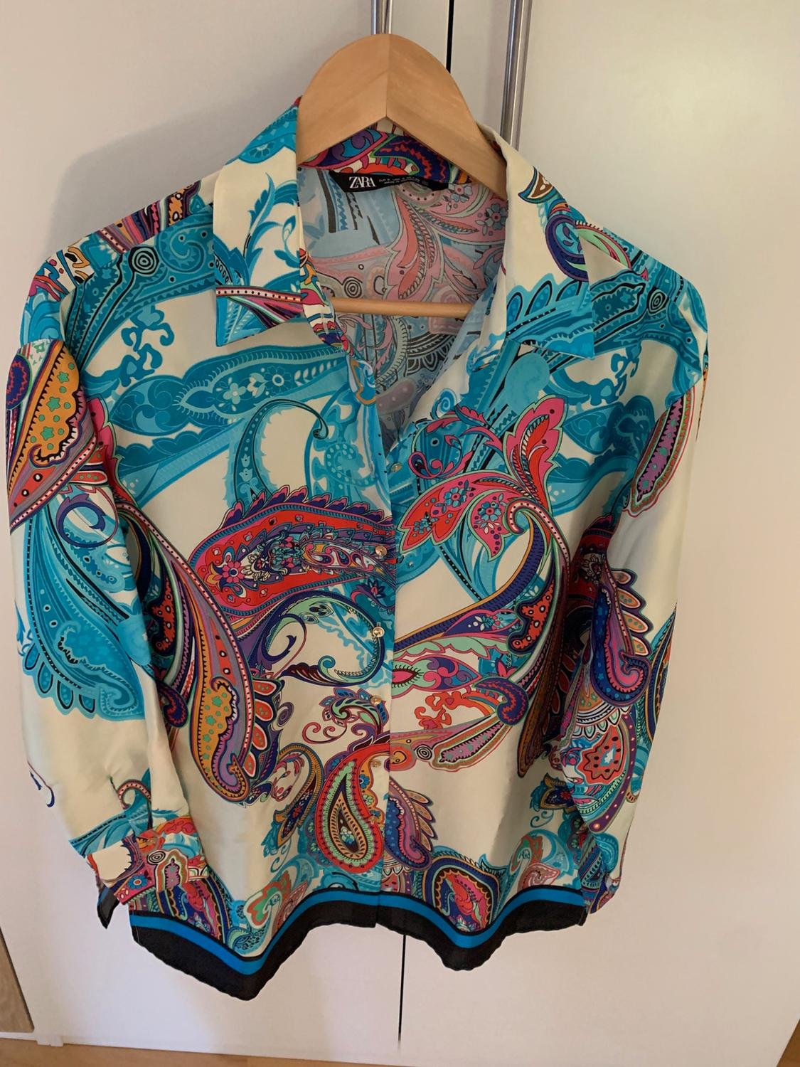 Button-up shirt with a multicoloured paisley print in shades of blue and pink, hanging on a wooden hanger. Label with the text ZARA visible.