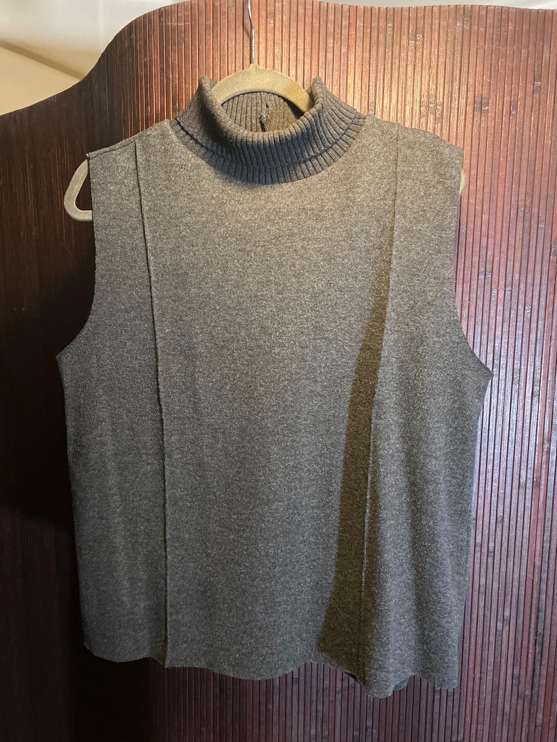 Sleeveless gray knit turtleneck vest, hanging on a hanger in front of a brown wood-paneled background.