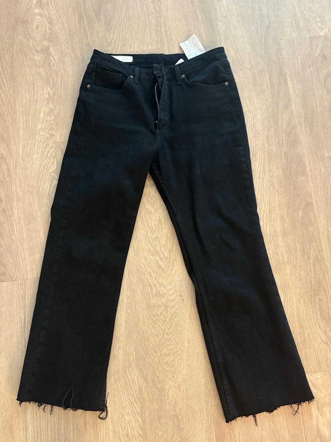 Black jeans with a frayed hem and a slightly open zipper, laid out on a light wood floor.