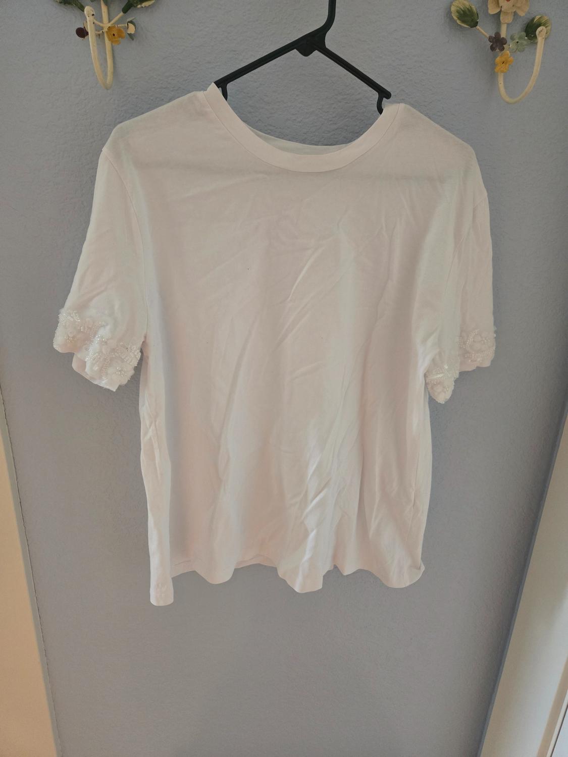 White short-sleeved T-shirt with lace details on the sleeves, hanging on a black hanger against a grey wall.
