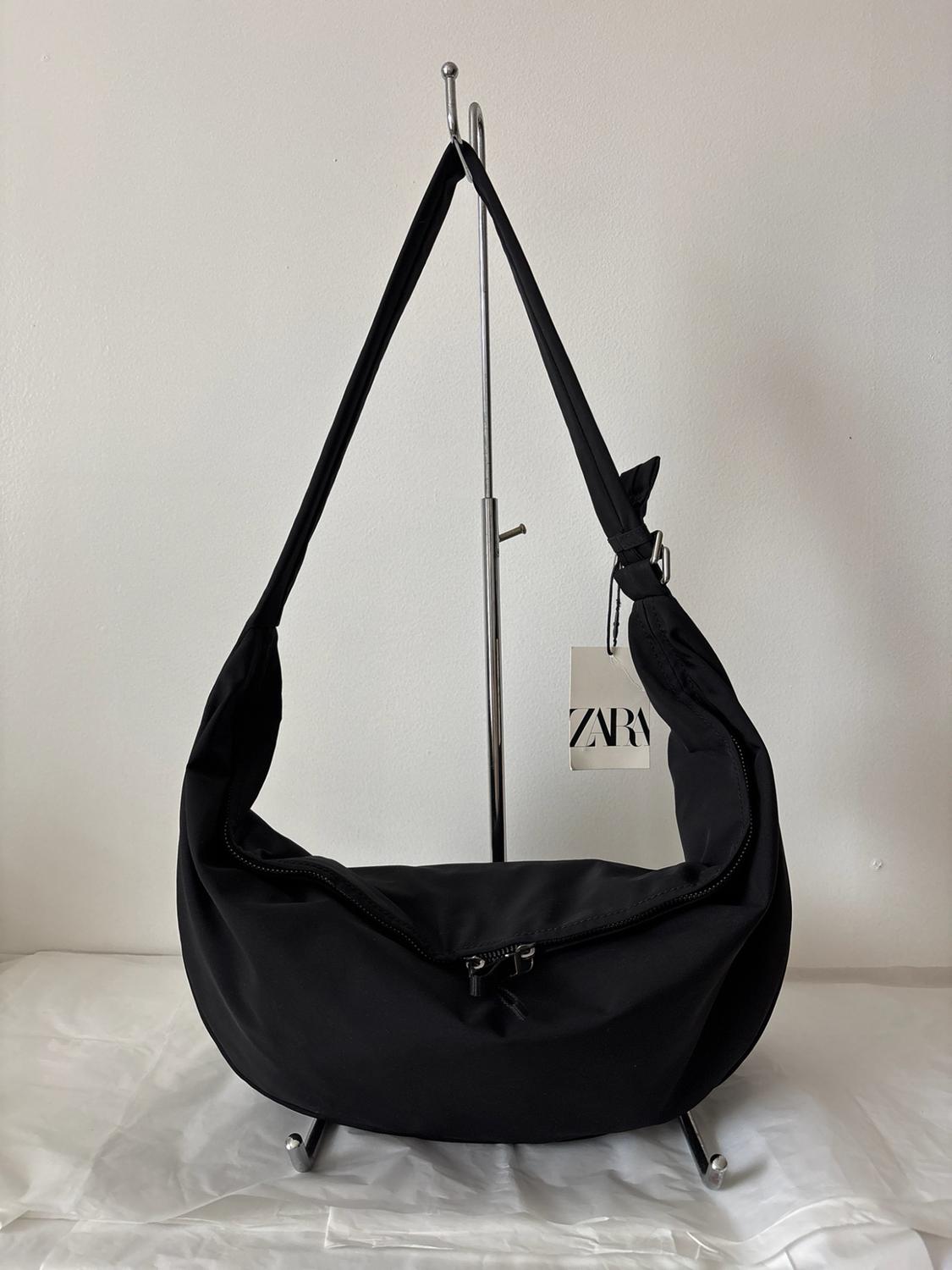 Black zipped handbag hanging on a metal stand against a minimalist white background. Visible label with the text ZARA.