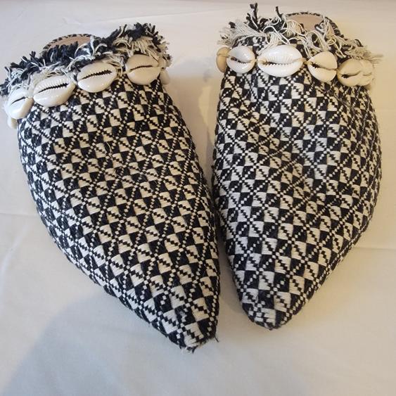 Black and white woven shoes with a geometric pattern, decorated with shells and fringe on the top, on a white background.