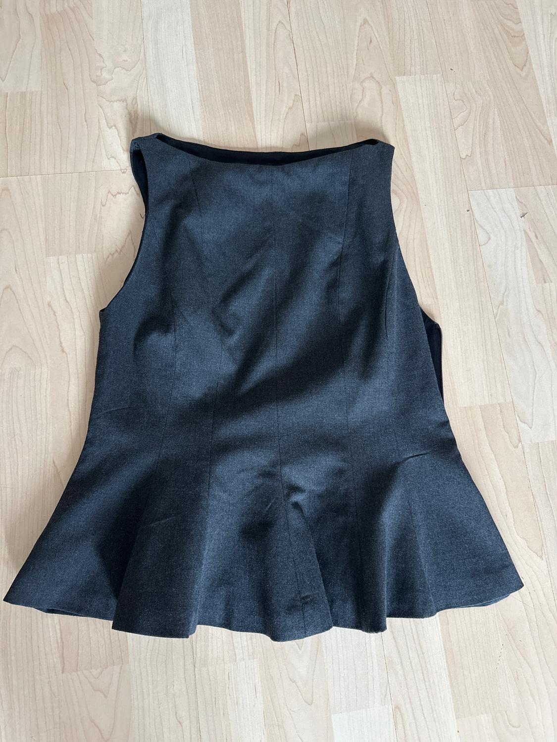 Dark grey sleeveless waistcoat with a flared design at the bottom, on a light wooden floor.