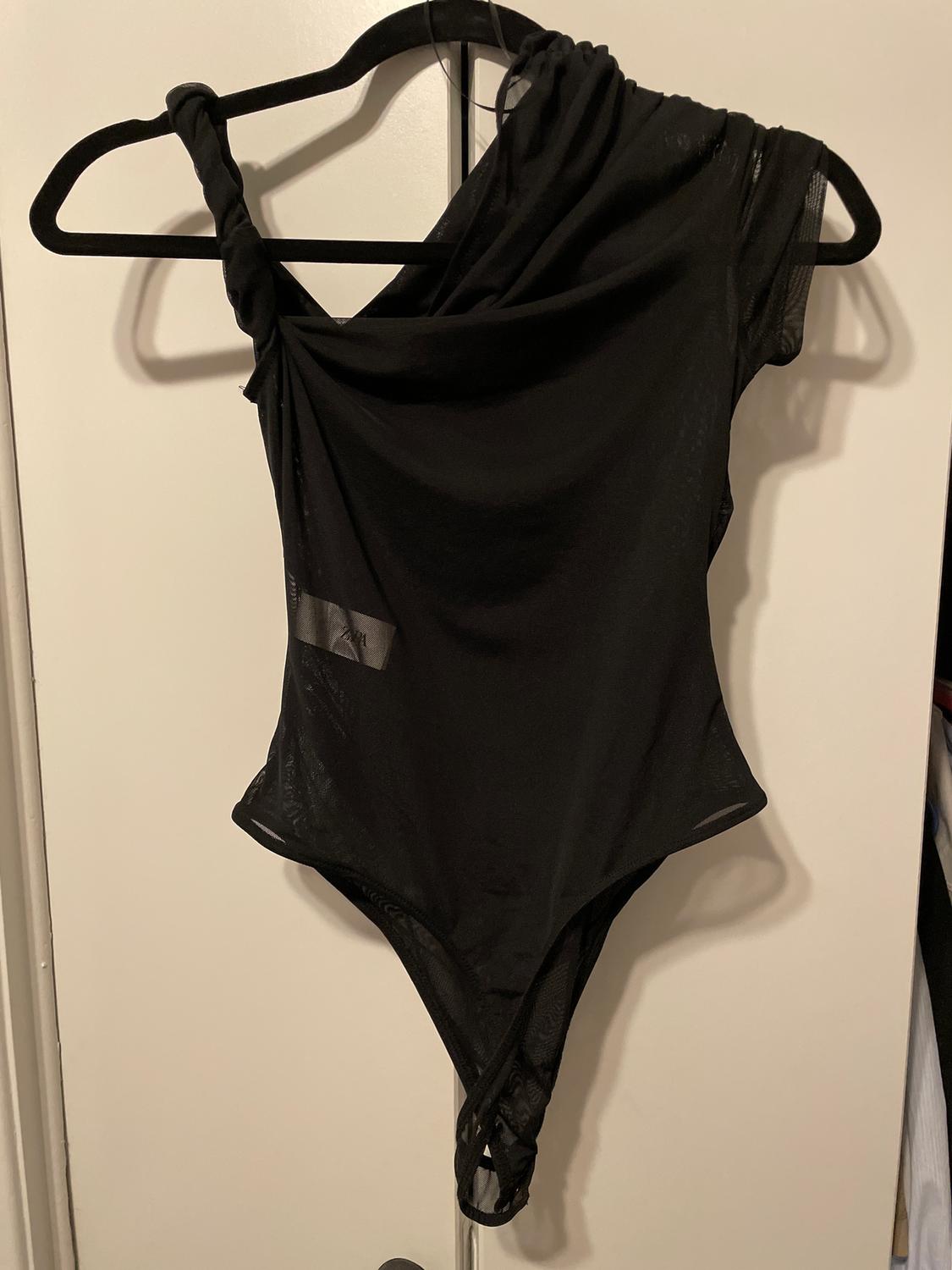 Asymmetrical black bodysuit hanging on a black hanger over a white door. Lightweight fabric with a sleek, modern design.