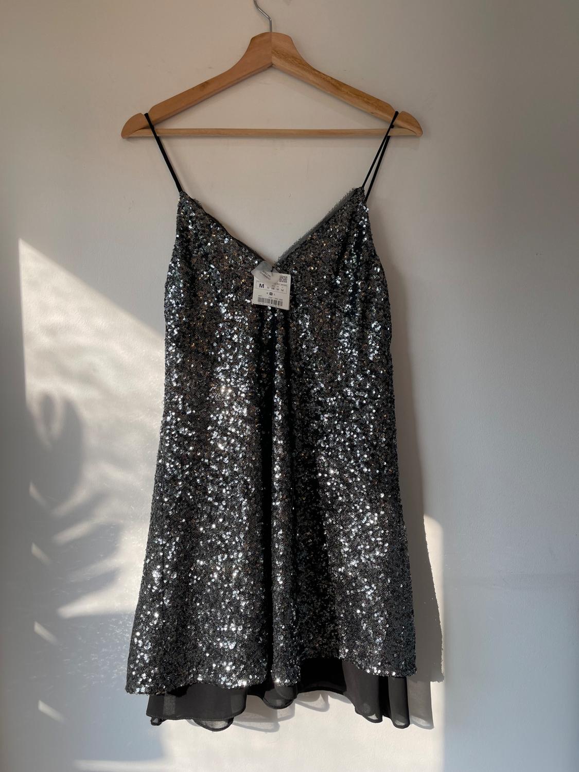 Silver sequin dress hanging on a wooden hanger, with thin straps and visible tag, illuminated by natural light.