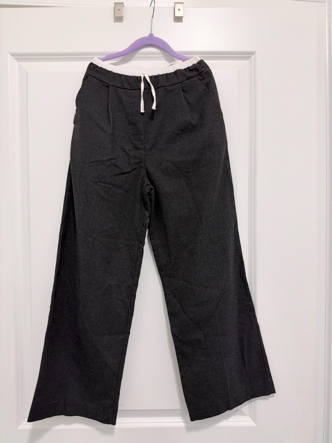 Dark gray wide-leg pants with a white elastic waistband and adjustable drawstring, hanging on a lilac hanger on a white door.