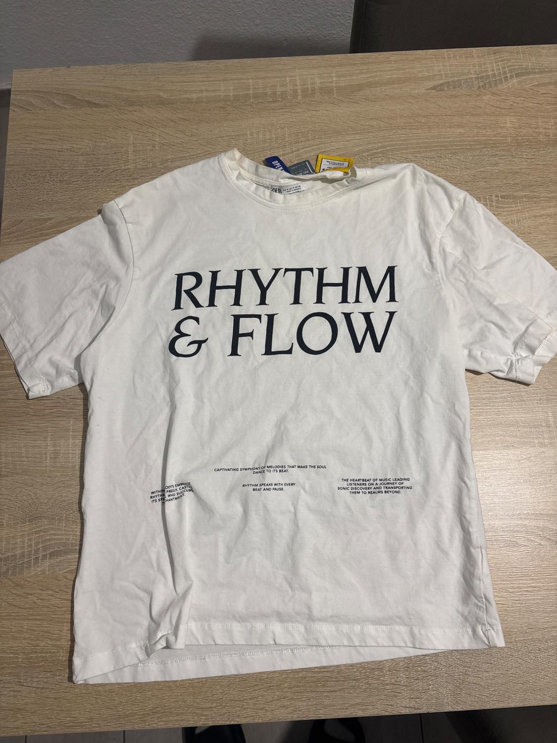 White T-shirt on a wooden table with the text "RHYTHM & FLOW" on the front. Labels visible at the neck. Small additional text at the bottom.