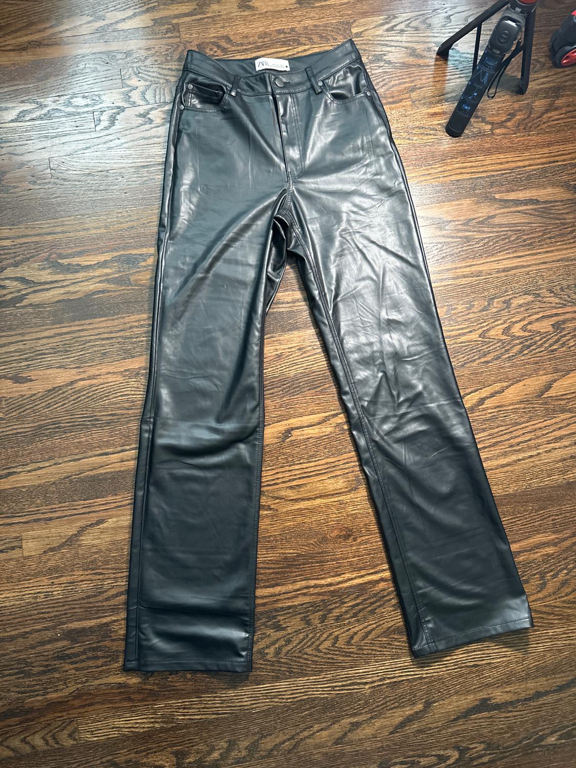 Black leather pants on a wooden floor. Visible tag at the waist. Part of a tripod in the upper right corner.