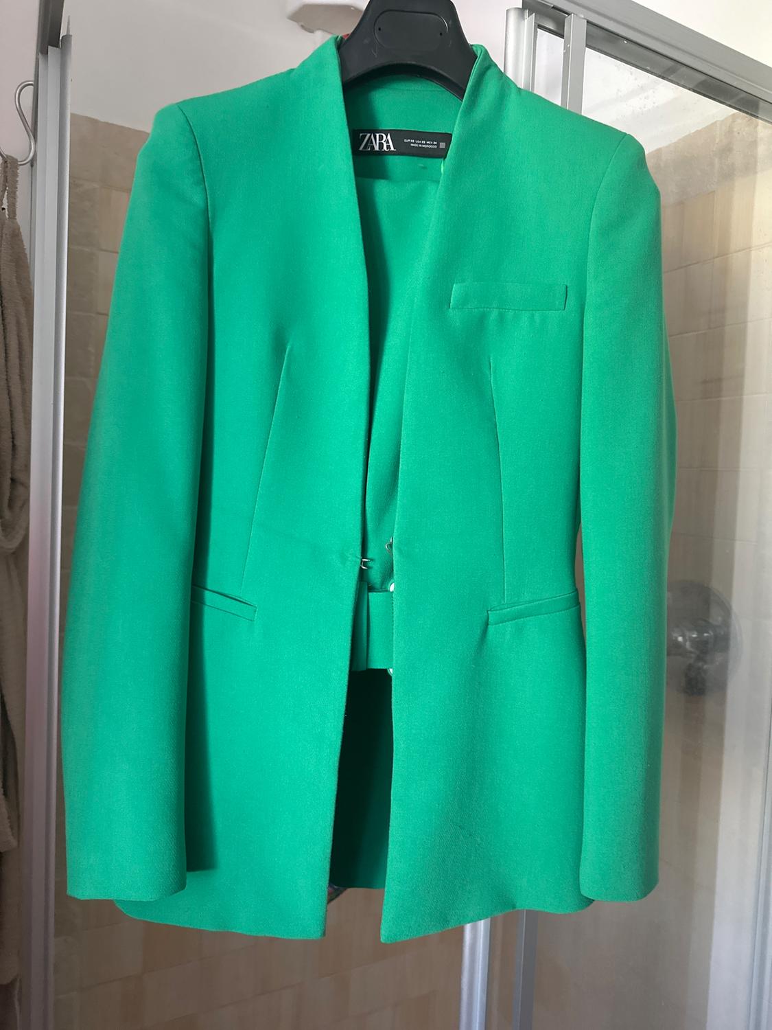 Green jacket hanging on a black hanger inside a bathroom. Visible label: "ZARA". Tiled background with a beige towel.