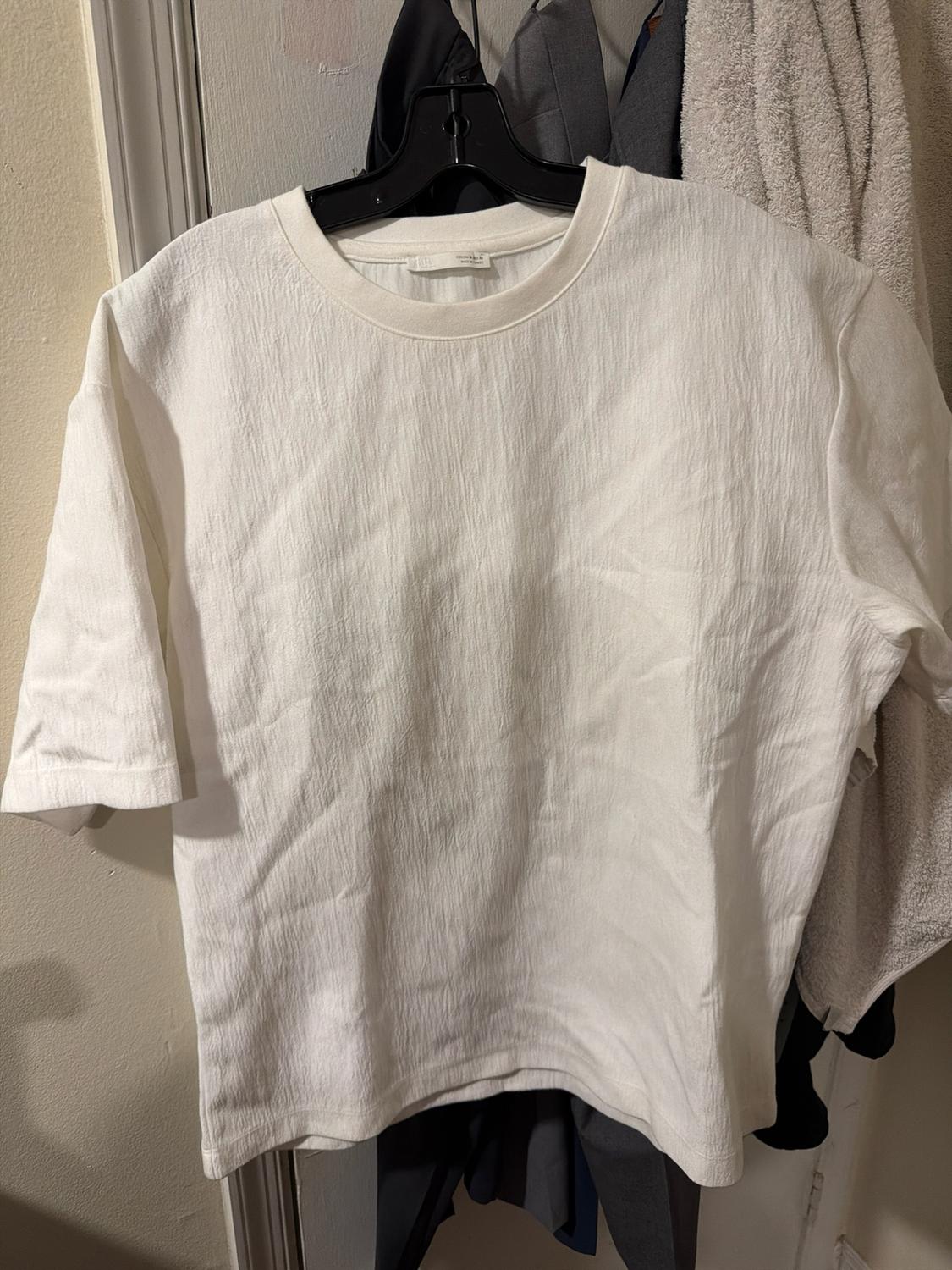 White short-sleeved textured T-shirt, hanging on a black hanger in front of a door with other clothes in the background.