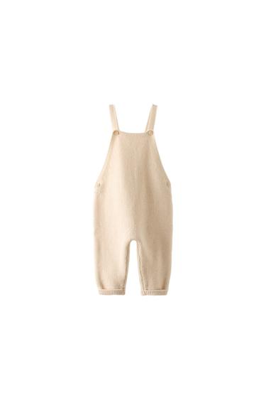 Front view of beige knitted dungarees with straps and buttons.