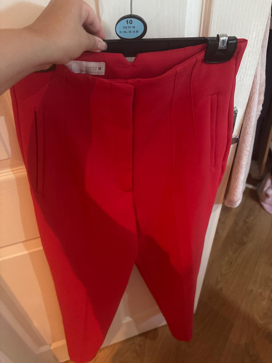 Hand holding red trousers on a hanger in front of a white door. Text: 10 ES/PT 38 D/NL/B/A 36.